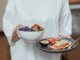 Person holding a bowl of fresh berries and a plate with avocado toast, displaying a healthy breakfast spread.